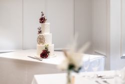 Three-tier white wedding cake adorned with deep red flowers and a cake topper, displayed on a white tablecloth.