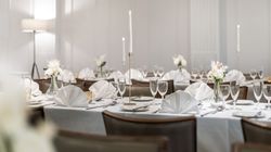 Elegant dining table set for a formal event with white tablecloths, folded napkins, glassware, white floral centerpieces, and tall lit candles.