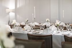 Elegant dining table set for a formal event with white tablecloths, folded napkins, glassware, white floral centerpieces, and tall lit candles.