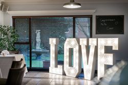 Large illuminated LOVE letters at an indoor event, with a dining table on the left and a congratulatory chalkboard sign in the background.