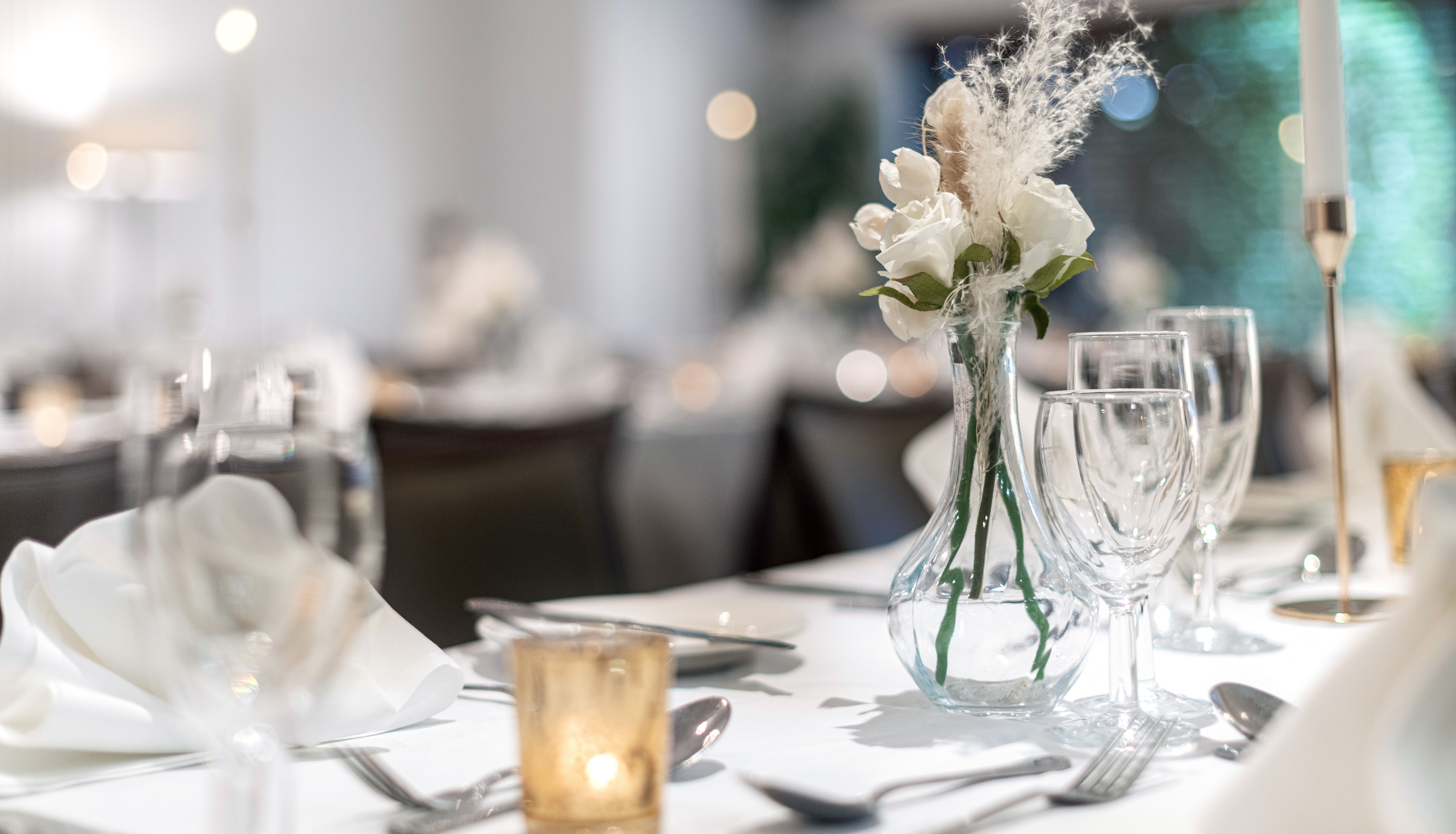 Elegant table setting with white napkins, clear glassware, a small vase of white flowers, a lit candle, and silverware, suggesting a formal event or wedding reception.