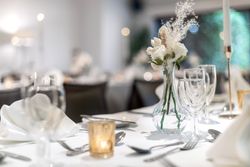 Elegant table setting with white napkins, clear glassware, a small vase of white flowers, a lit candle, and silverware, suggesting a formal event or wedding reception.