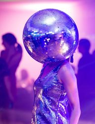Person wearing a large disco ball helmet and a sequined outfit at a party with purple lighting and people in the background.