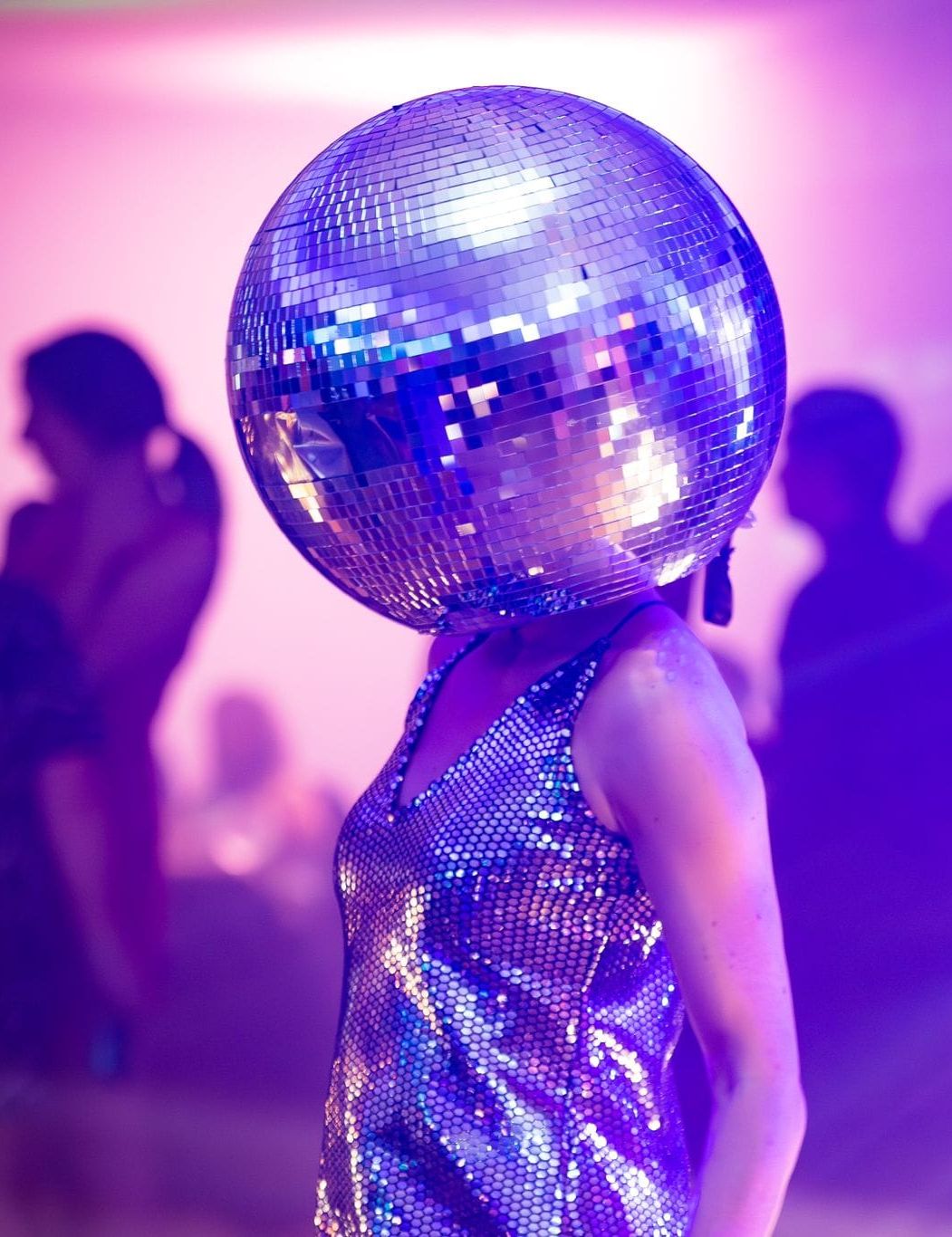 Person wearing a large disco ball helmet and a sequined outfit at a party with purple lighting and people in the background.