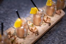 Mini mason jars of a pinkish beverage with orange garnishes and straws, served on a wooden board with grilled chicken skewers.