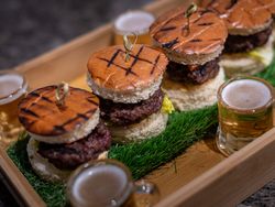 Tray of mini hamburgers with grilled buns, served with small glasses of beer, displayed on artificial grass.