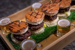 Tray of mini hamburgers with grilled buns, served with small glasses of beer, displayed on artificial grass.