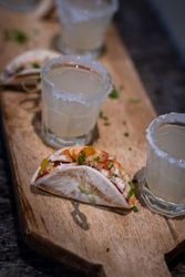 Close-up of a wooden serving board with small tacos filled with diced vegetables and meat, and glasses of a light-colored beverage with salted rims.