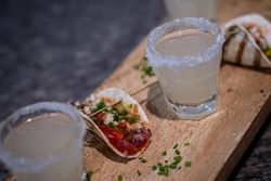 Close-up of two glasses with salted rims filled with a light-colored drink next to a small taco topped with diced vegetables and herbs, all placed on a wooden serving board.