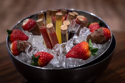 A metal bowl filled with ice, containing test tubes with colorful liquids sealed with corks, and fresh strawberries, some dipped in chocolate.