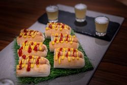 Several hot dogs topped with ketchup and mustard are arranged on a tray with artificial grass. In the background, there are three shot glasses filled with a foamy yellow beverage.
