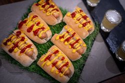 A group of six hot dogs in buns, topped with ketchup and mustard, arranged on a piece of artificial grass next to glasses of beer.