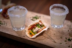 Two shot glasses with salted rims filled with a clear drink, placed beside a small taco with chicken, bell peppers, tomatoes, and green garnish on a wooden board.