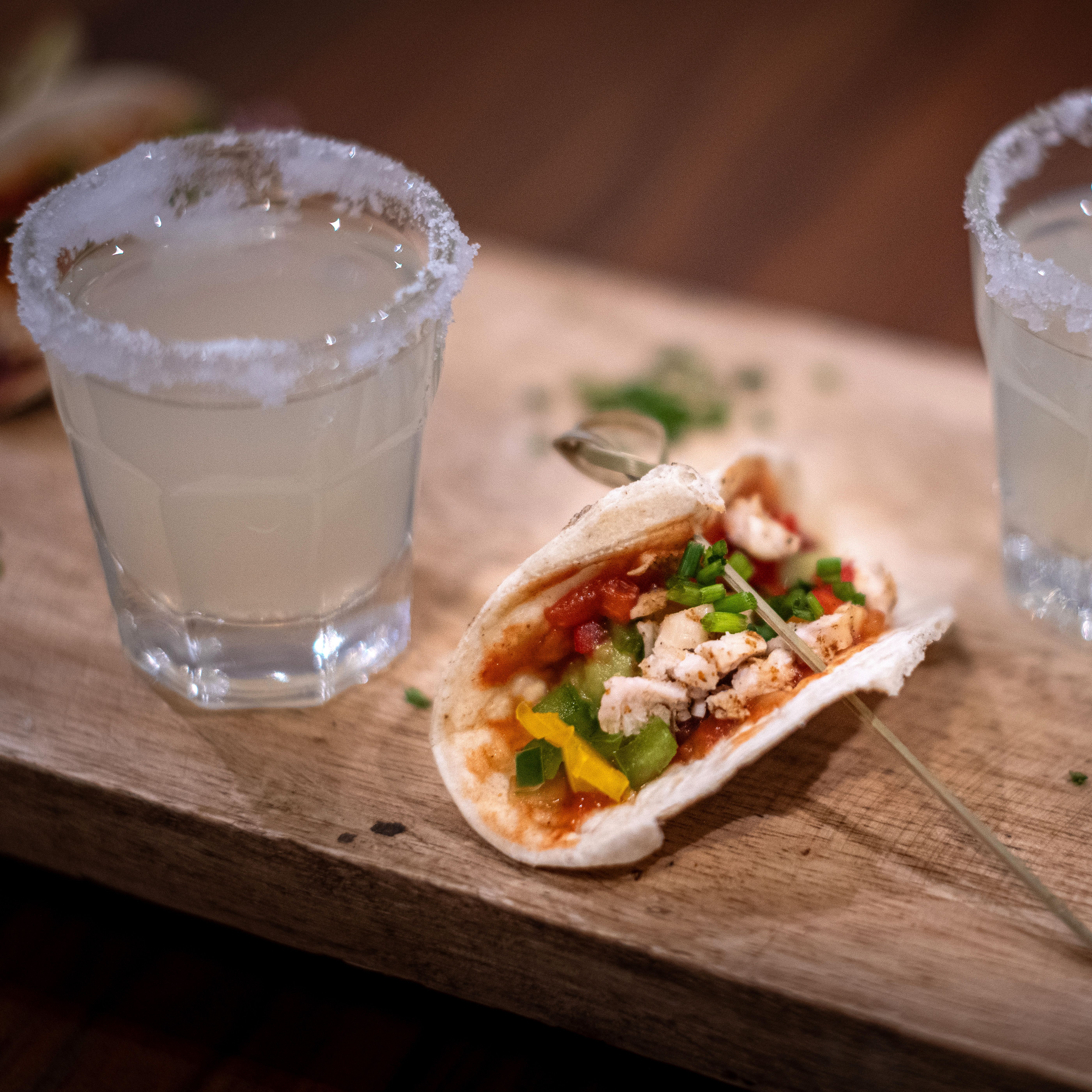 Two shot glasses with salted rims filled with a clear drink, placed beside a small taco with chicken, bell peppers, tomatoes, and green garnish on a wooden board.
