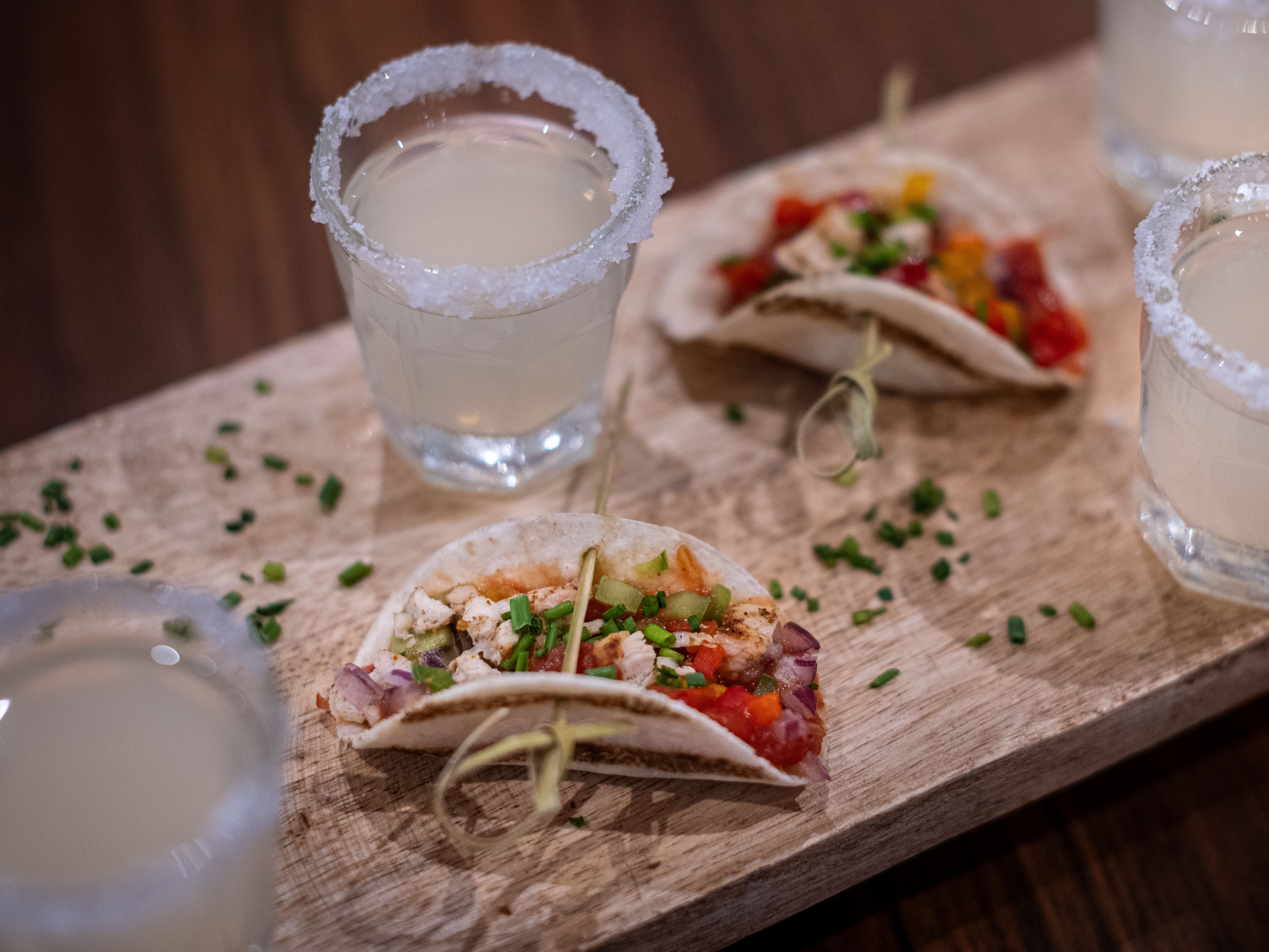 Mini tacos garnished with chopped chives served on a wooden board alongside glasses of a light drink with salted rims.