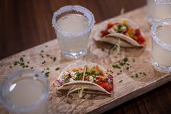 Mini tacos garnished with chopped chives served on a wooden board alongside glasses of a light drink with salted rims.