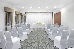 A small indoor wedding ceremony setup with rows of white covered chairs facing a table decorated with white flowers, in front of a backdrop of white curtains and soft lighting.