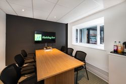 Modern meeting room with a wooden table, black chairs, a TV displaying the Holiday Inn logo on a dark accent wall, and refreshments on the side by a large window.