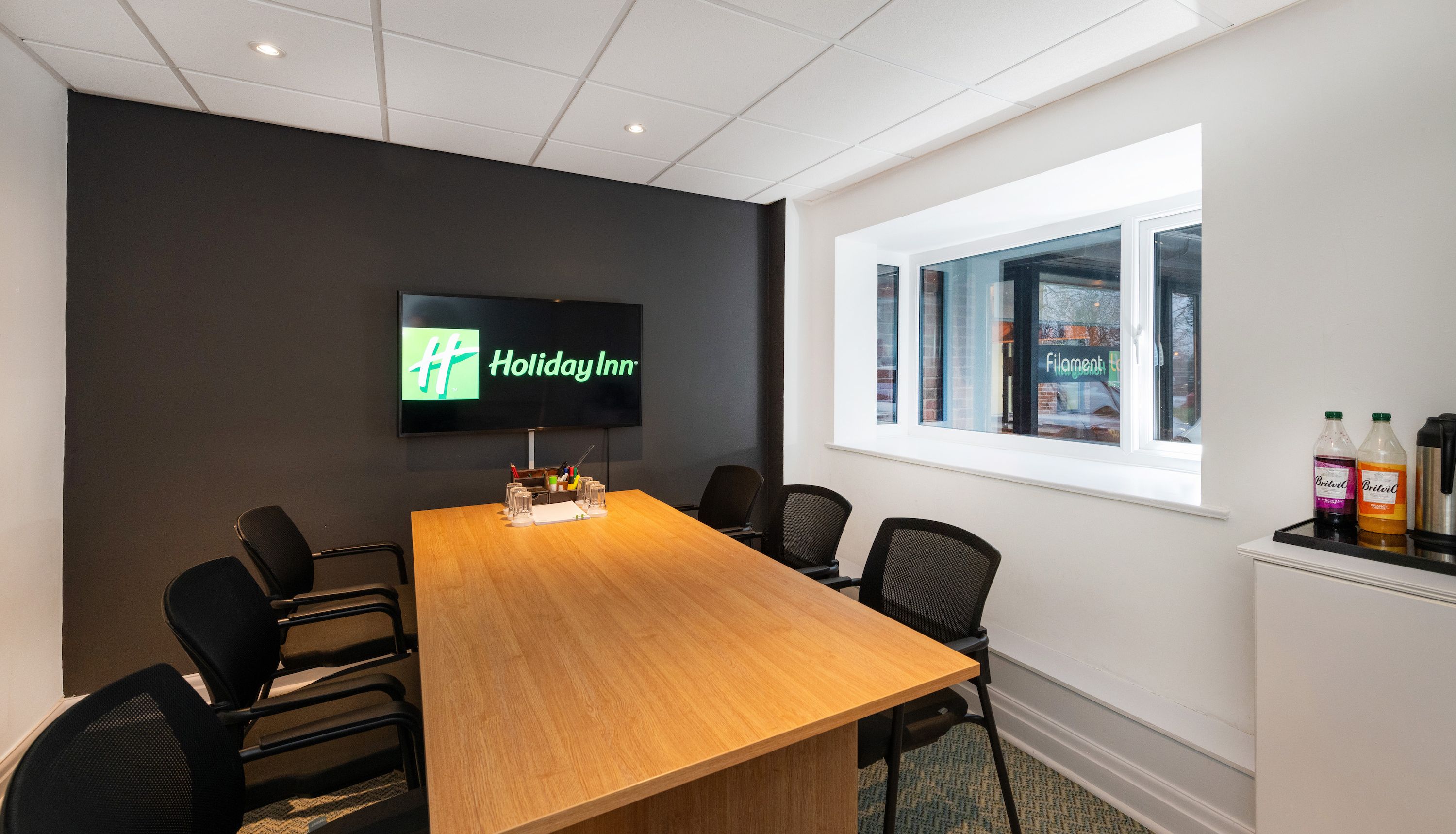 Modern meeting room with a wooden table, black chairs, a TV displaying the Holiday Inn logo on a dark accent wall, and refreshments on the side by a large window.