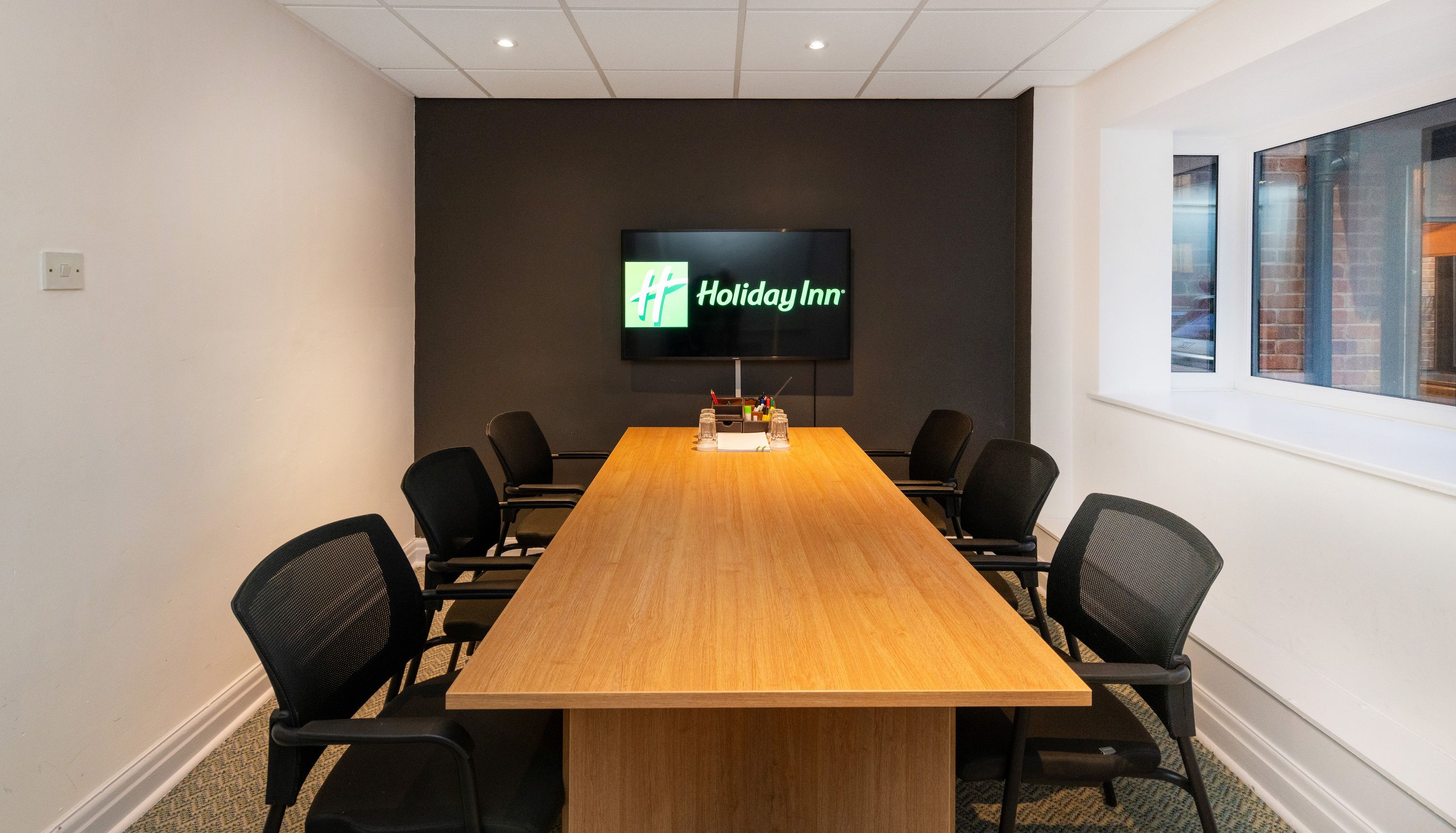 Modern conference room with a wooden table, six black office chairs, a television screen displaying the Holiday Inn logo, and large windows on the right side.