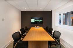 Modern conference room with a wooden table, six black office chairs, a television screen displaying the Holiday Inn logo, and large windows on the right side.