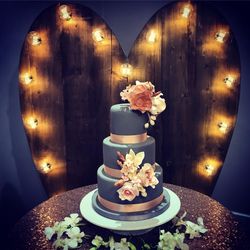 Elegant three-tiered cake with gray fondant, decorated with pink and white sugar flowers, set against a large heart-shaped wooden backdrop with string lights.