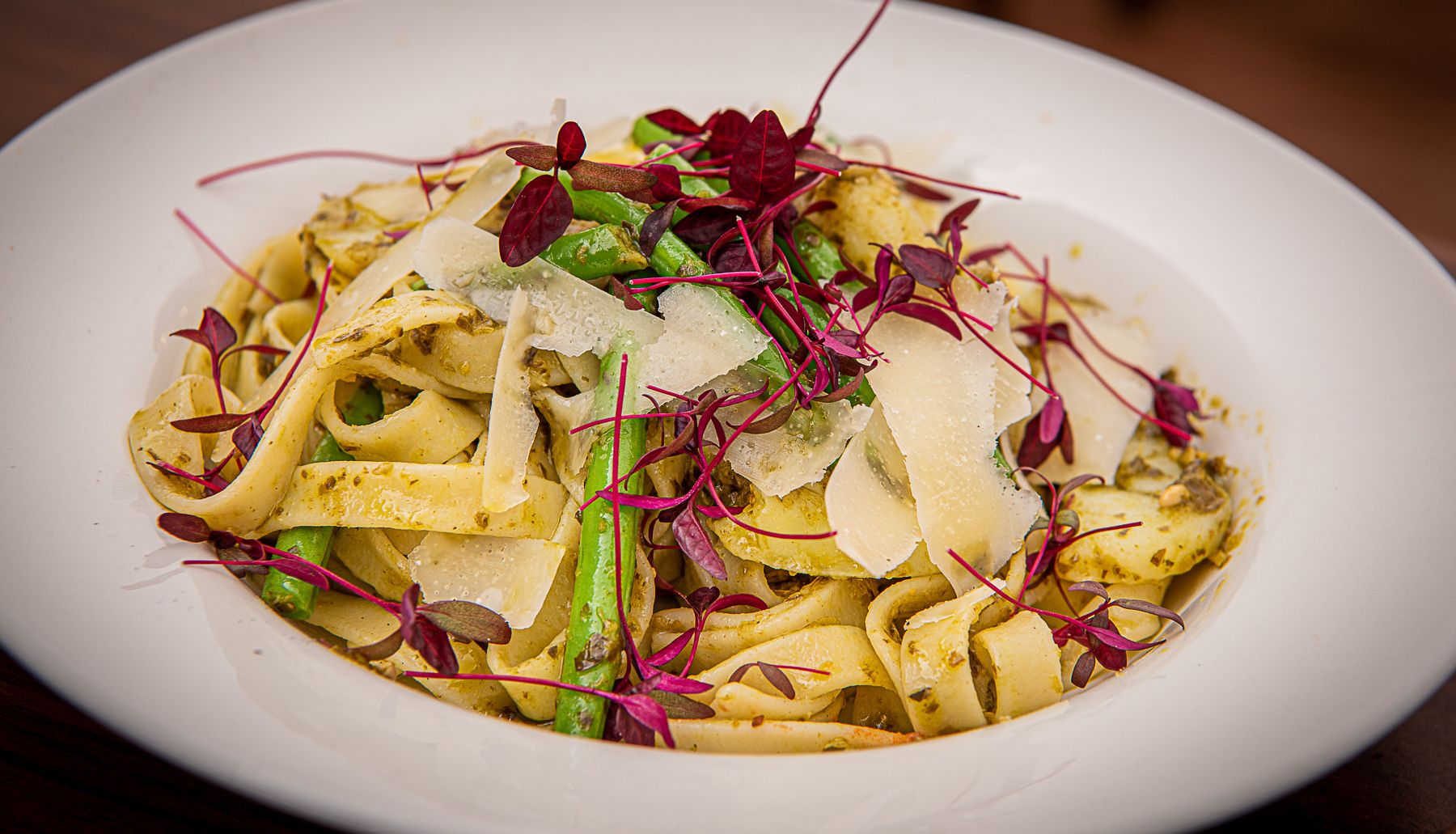 A plate of tagliatelle pasta with green beans, potato pieces, pesto sauce, parmesan cheese shavings, and microgreens garnish.