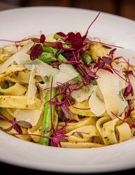 A plate of tagliatelle pasta with green beans, potato pieces, pesto sauce, parmesan cheese shavings, and microgreens garnish.