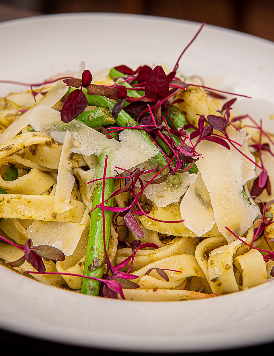 A plate of tagliatelle pasta with green beans, potato pieces, pesto sauce, parmesan cheese shavings, and microgreens garnish.