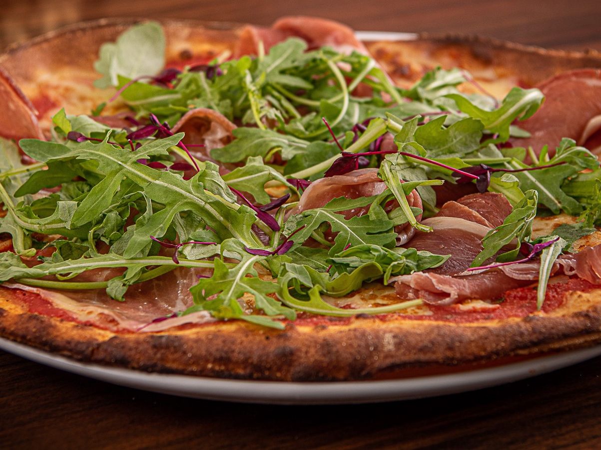 A close-up of a thin crust pizza topped with arugula, prosciutto, and tomato sauce on a white plate.