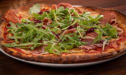 A close-up of a thin crust pizza topped with arugula, prosciutto, and tomato sauce on a white plate.
