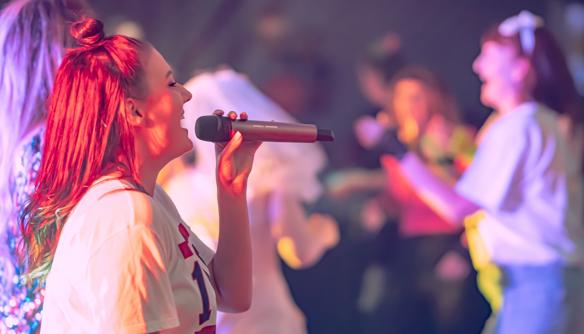 Woman singing into a microphone on stage with colorful lighting and blurred audience in the background.