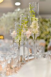 Elegant table centerpiece with tall glass vases holding white flowers and floating candles, set on a sparkling table runner in a formal event setting.
