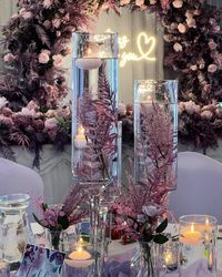 Elegant table centerpiece with tall glass vases filled with water, floating candles, and purple floral arrangements, set against a backdrop of flowers and a neon sign with a heart.