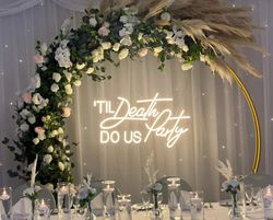 Wedding reception decoration with floral arch and neon sign reading 'TIL DEATH DO US Party'