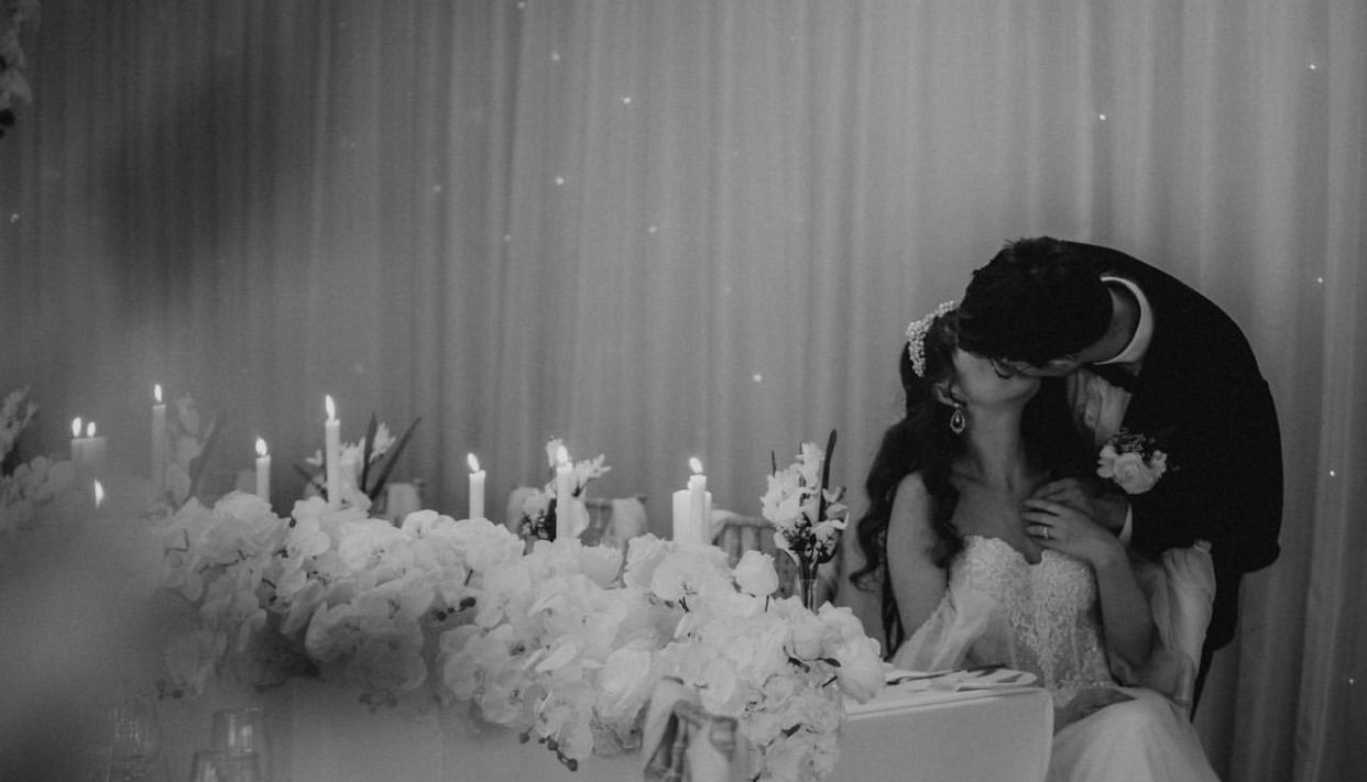 Black and white photo of a bride and groom kissing at their wedding reception, seated at a table decorated with flowers and lit candles.