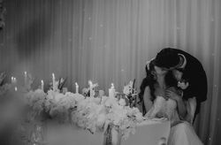 Black and white photo of a bride and groom kissing at their wedding reception, seated at a table decorated with flowers and lit candles.