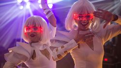 Two women in futuristic white costumes and white wigs wearing glowing red visor glasses at a party or event.