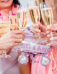 Group of women toasting with champagne glasses at a baby shower, holding a pink 'It's a Girl!' balloon.