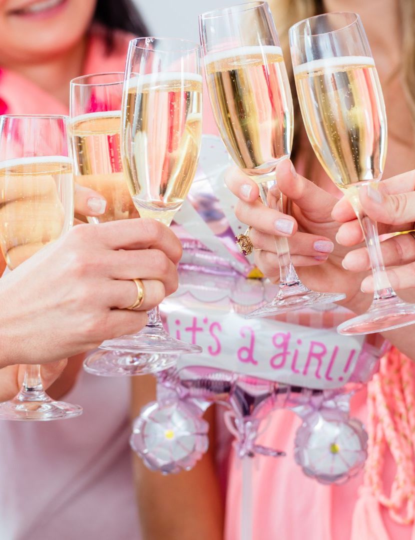 Group of women toasting with champagne glasses at a baby shower, holding a pink 'It's a Girl!' balloon.