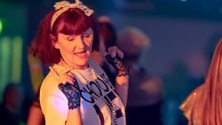 Woman dressed in 1980s style, wearing a white t-shirt with bold black text, lace gloves, a pearl necklace, and a white hair bow, dancing at a party with colorful lighting.