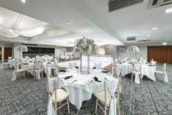 Elegant wedding reception hall decorated in white with round tables, floral centerpieces, draped chairs, and a large illuminated 'LOVE' sign near the dance floor.