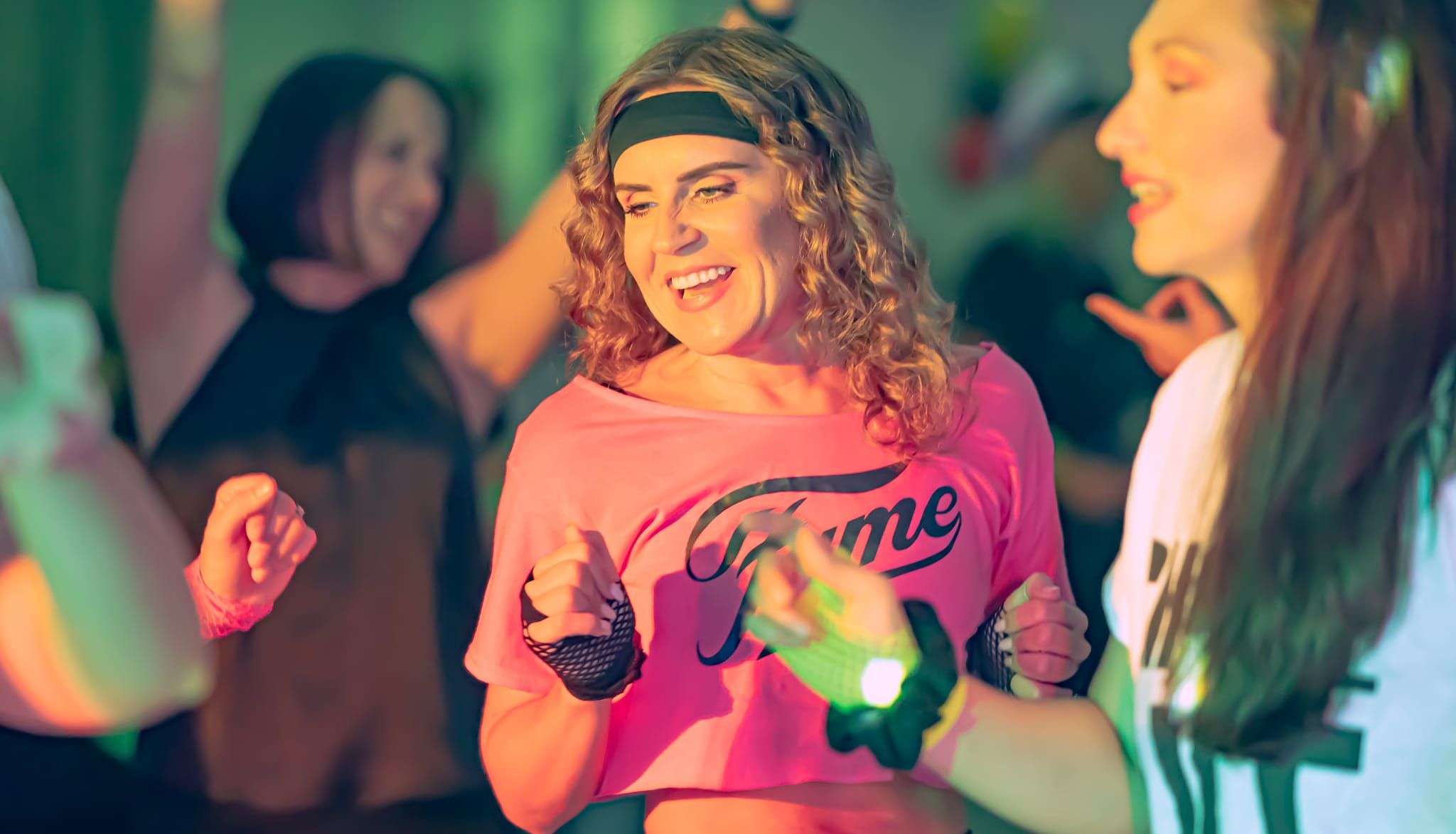 Women dressed in 1980s-style workout clothes dancing and smiling at a retro-themed party or fitness class.