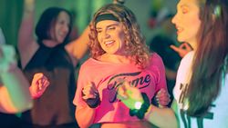Women dressed in 1980s-style workout clothes dancing and smiling at a retro-themed party or fitness class.