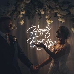 Bride and groom holding hands and smiling in front of a floral arch with a neon sign that reads 'Happily Ever After'.