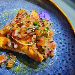A close-up of a slice of toasted bread topped with finely chopped tomatoes, onions, herbs, and microgreens, served on a blue ceramic plate.
