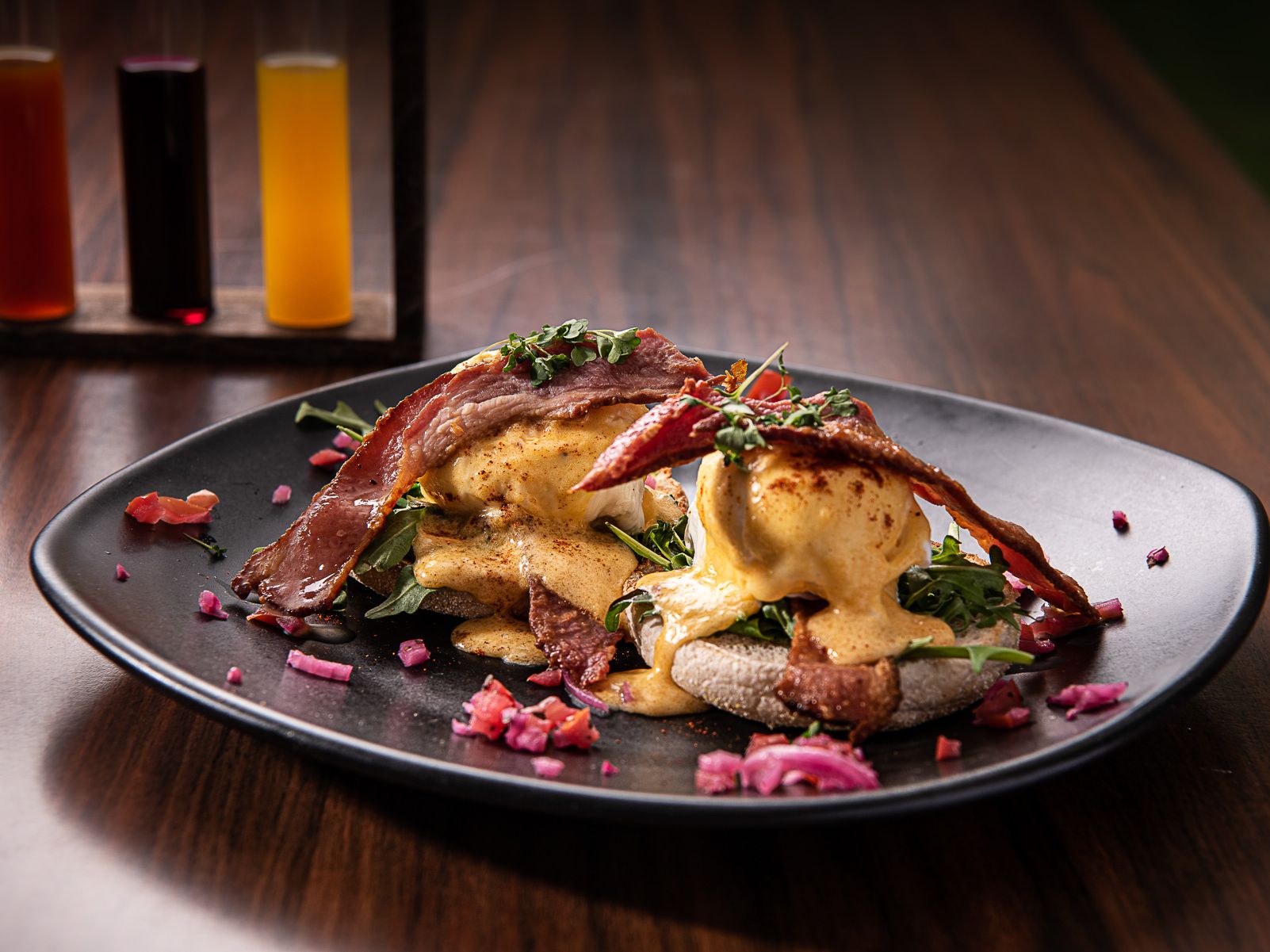 Eggs Benedict with crispy bacon, poached eggs, hollandaise sauce, and greens served on an English muffin, garnished with microgreens and pickled vegetables on a black plate.