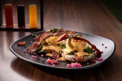 Eggs Benedict with crispy bacon, poached eggs, hollandaise sauce, and greens served on an English muffin, garnished with microgreens and pickled vegetables on a black plate.