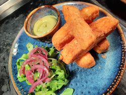 A blue plate with several golden, crispy fried sticks, a small bowl of green dipping sauce, and a garnish of fresh arugula topped with pickled red onions.