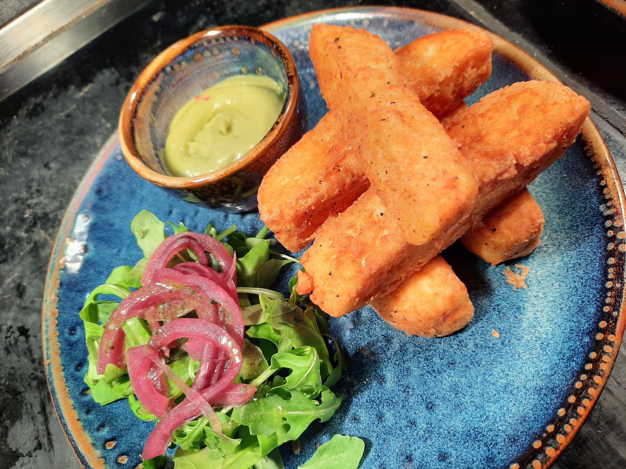 A blue plate with several golden, crispy fried sticks, a small bowl of green dipping sauce, and a garnish of fresh arugula topped with pickled red onions.